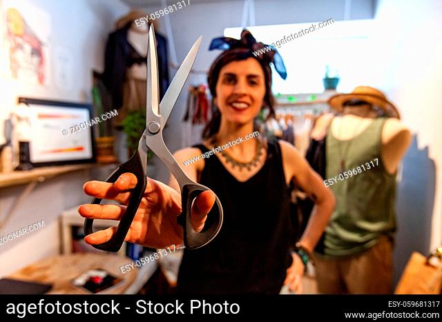 A trendy seamstress is viewed from the front holding a pair of crafting scissors inside an atelier. Creative person showing tools of trade