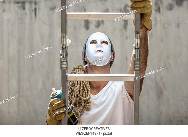 Man wearing a mask climbing a ladder with rope and a can of spray paint