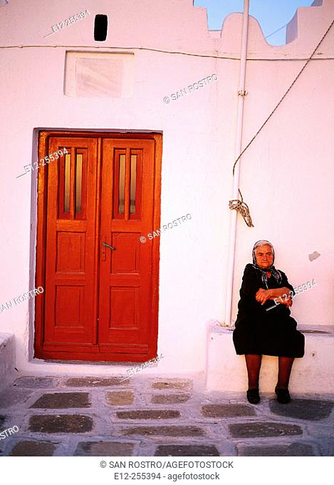 Greece, Cyclades Islands, Mykonos Senior lady looking having a rest in front of her door in a white washed street