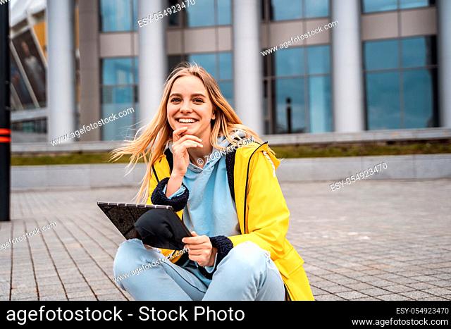 A beautiful blonde girl sits on a street and uses a tablet to communicate with friends or for the Internet or reading a book or another