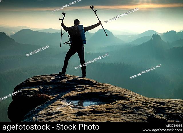 Silhouette of happy man with arms outstretched holding crutches standing by mountain valley during sunset. Personal motivation