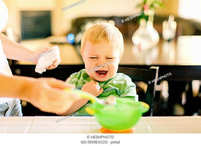 Mother helping young son eat breakfast, son crying, mid section