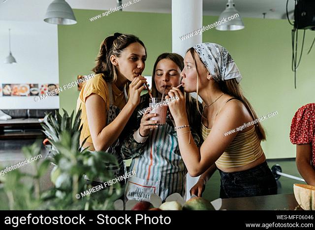 Teenage girls standing in kitchen sharing fresh fruit smoothie with drinking straws