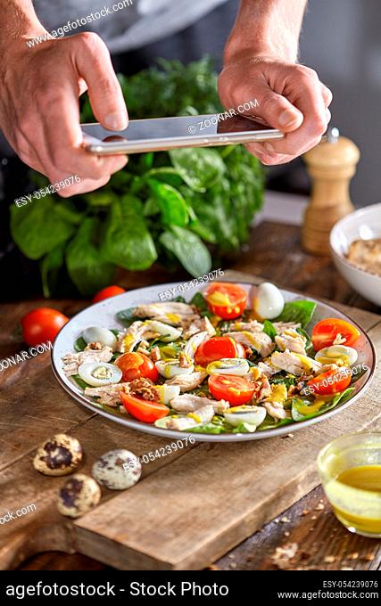 Delicious healthy salad from organic ingredients in a plate with mens hand making the photo by smartphon on a wooden background