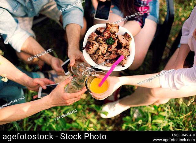Friendly group of people sitting in circle. Group of friends clinking their drinks and bowl of fried meat while sitting outside on grass