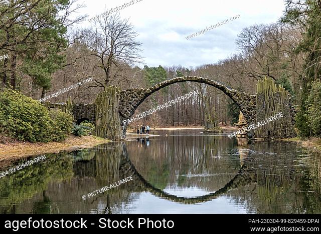 04 March 2023, Brandenburg, Kromlau: The Rakotz Bridge made of basalt stones in the Kromlau Rhododendron Park is reflected in the Rakotz Lake in overcast...