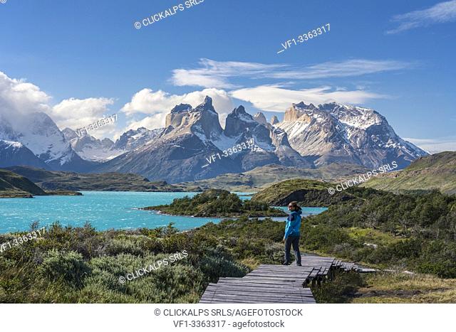 Woman standing on a boardwalk with Lake Pehoé and Paine Horns in the background, on a windy summer day. Torres del Paine National Park