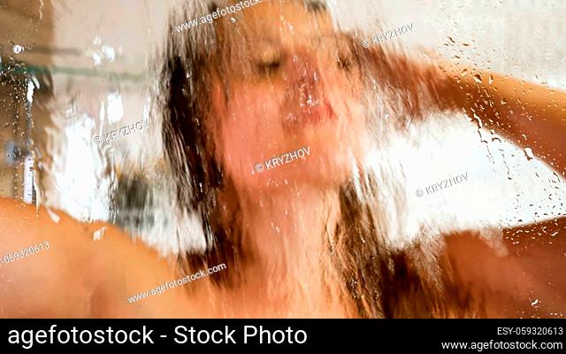 View through wet glass on beautiful brunette woman relaxing in shower and washing long hair. Concept of female beauty, body care and healthcare