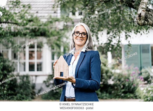 Portrait of smiling businesswoman with piece of wood shaped like a house standing in the garden