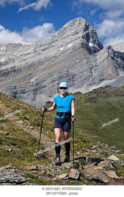 Female Hiker With Hiking Poles On A Mountain Trail With Mountain Peak In The Background And Blue Sky With Clouds, Alberta Canada
