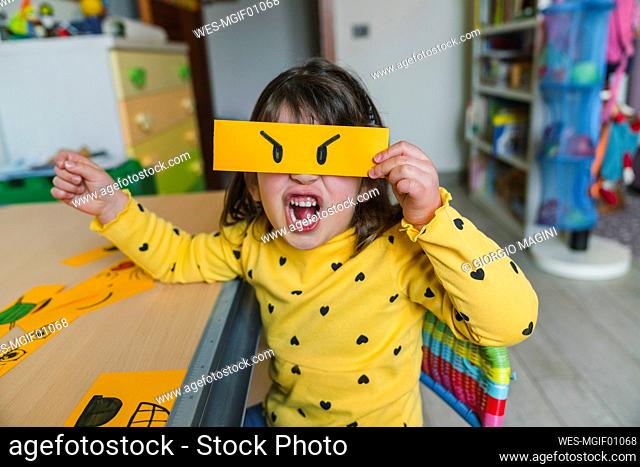 Girl making angry face while holding emoticon at home