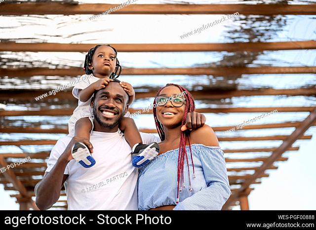 Smiling man carrying daughter on shoulders by woman under roofing