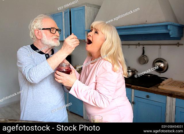 Cute old man feeding his wife jam from spoon. Elderly man wearing glasses and blue shirt feeding sweet old lady red jelly from jar