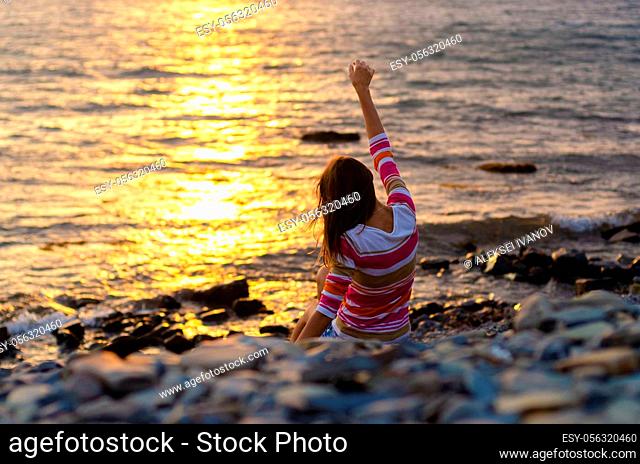 A girl sits on the seashore and throws stones into the water, raised her hands, evening, sunset