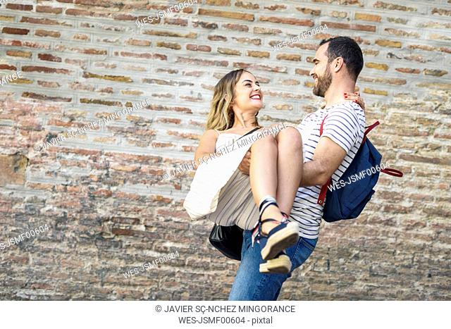 Happy man with backpack carrying girlfriend at brick wall