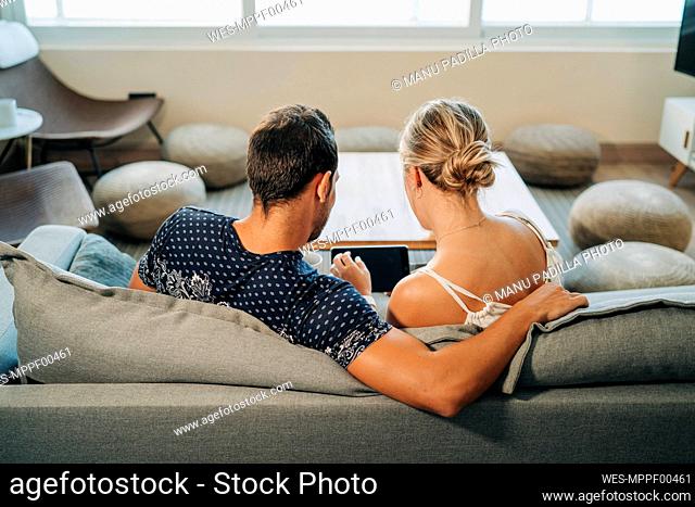 Rear view of couple sitting on couch in living room sharing a tablet