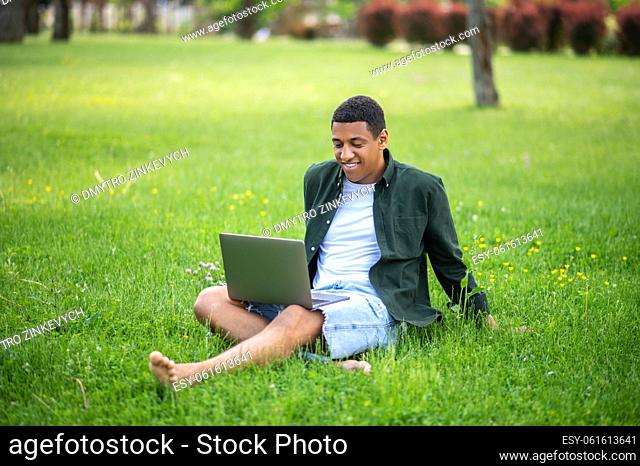 Good time. Smiling guy looking with interest at laptop sitting on grass in park on warm day