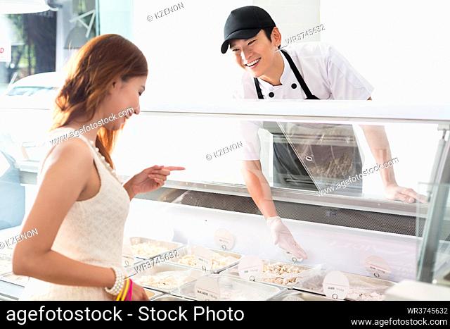 Smiling young Asian worker assisting a customer in a delicatessen as she selects food from a glass counter in the store