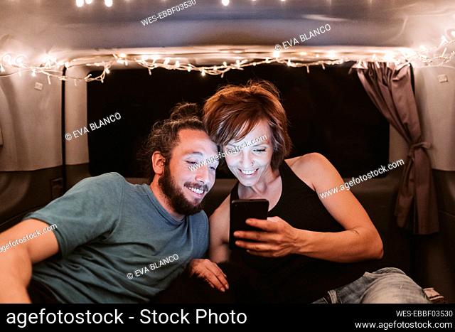 Smiling couple using mobile phone in illuminated van at night
