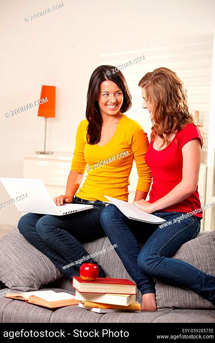 Teen girls sitting on couch at home with books and laptop, learning smiling at each other