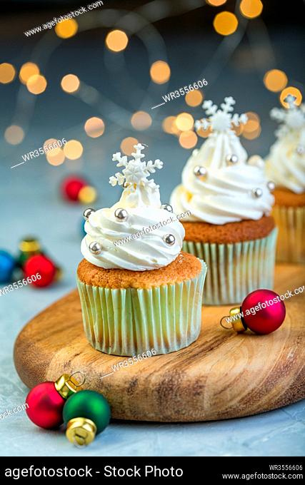 Cupcakes with vanilla cream, snowflakes and colorful Christmas balls on a wooden serving board on a blurred background of lights, selective focus