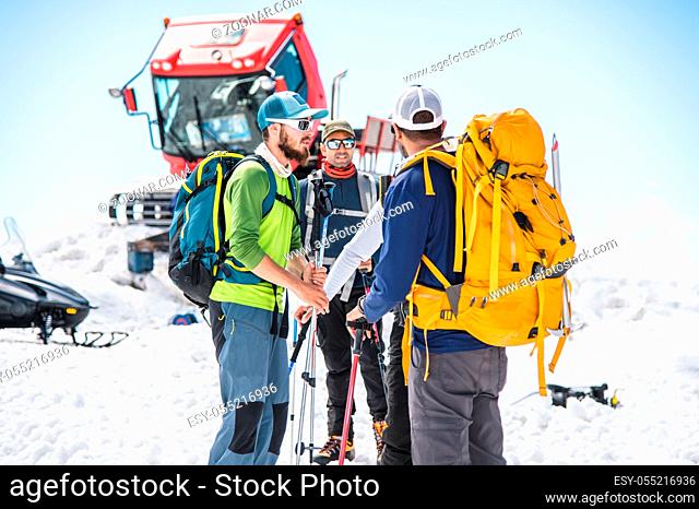 A team of climbers led by a guide discusses the upcoming climb against the background of a snow cat and mountains behind clouds