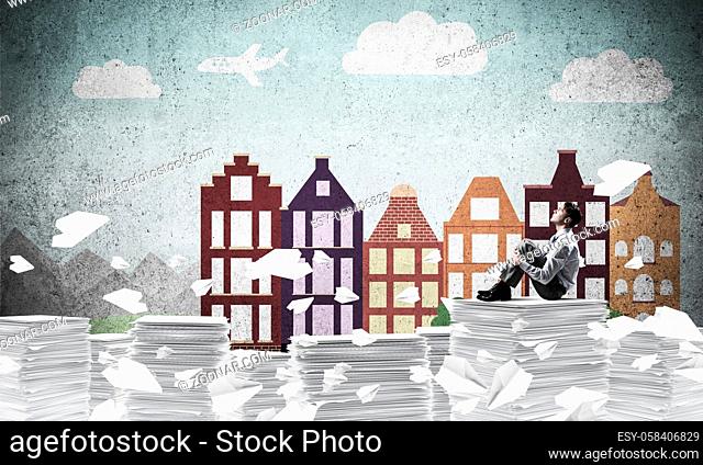 Thoughtful businessman looking away while sitting among flying paper planes with drawn cityscape on background. Mixed media
