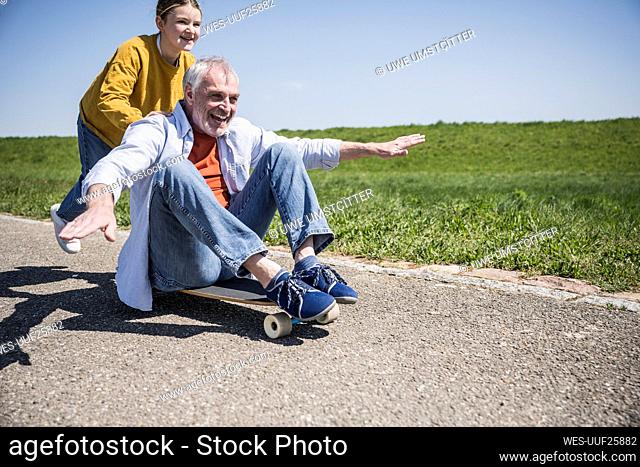 Happy girl pushing grandfather sitting with arms outstretched on skateboard