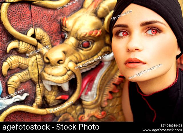 Fashion closeup portrait of young brunette woman with black hat and red eyes over golden dragon on the wall background