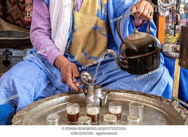 Berber man wearing turban and deraa pours tea from teapot into glasses for traditional Moroccan tea ceremony, Tighmert Oasis, Morocco