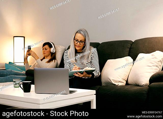 Mother with laptop writing in book while sitting with daughter in background at home