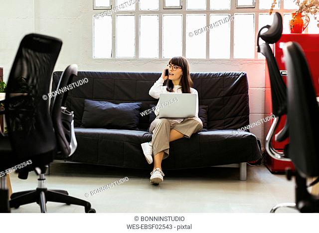 Young woman sitting on couch in office using cell phone and laptop