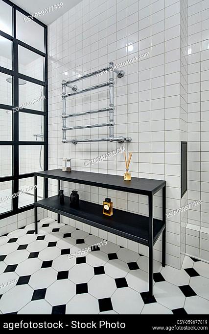 Stylish bathroom with walls of white tiles. There is shower with black partition of glass squares, black rack with accessories and chromed heated towel rail