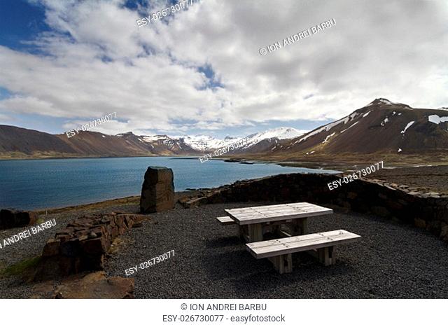 Horizontal panorama of a wooden resting bench next to a lake and mountains covered in snow in Iceland