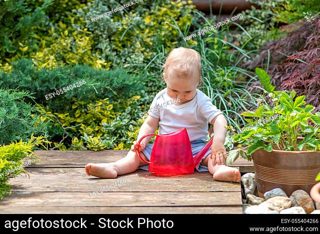 Cool little baby boy is sitting in the garden holding garden watering can