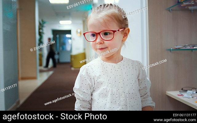 Adorable child blonde girl in ophthalmology clinic plays in hall near glasses store, close up