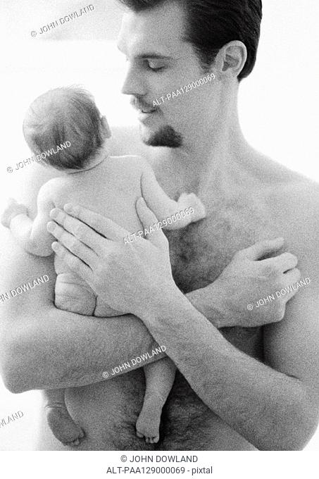 Topless father holding infant, patting infant's back, b&w