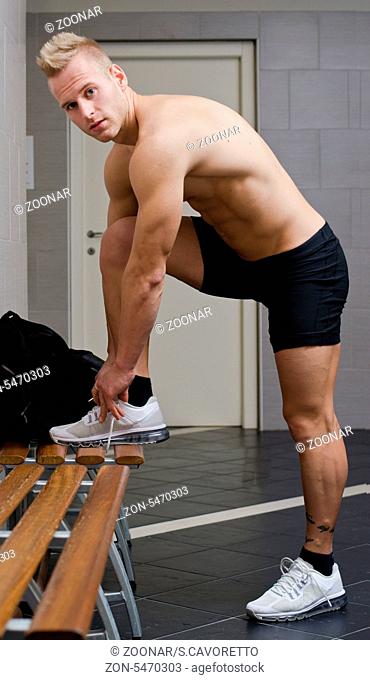 Handsome muscular athlete in locker room tying sneakers, shirtless, looking at camera