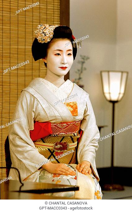 PORTRAIT OF A MAIKO APPRENTICE GEISHA IN A KIMONO OBEDE, WEARING A CHIGNON HIGH ON HER HEAD IN THE FORM OF A PEACH WARESHINOBU ADORNED WITH A RED SILK...