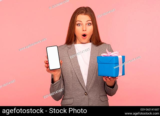Unbelievable bonus for mobile user, online shopping application. Portrait of surprised businesswoman in suit jacket holding cellphone and gift box