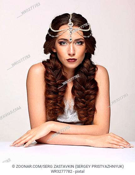 Beauty portrait of young woman in pearl diadem. Brunette girl with braids hairdo and day female makeup sit at white table on gray background