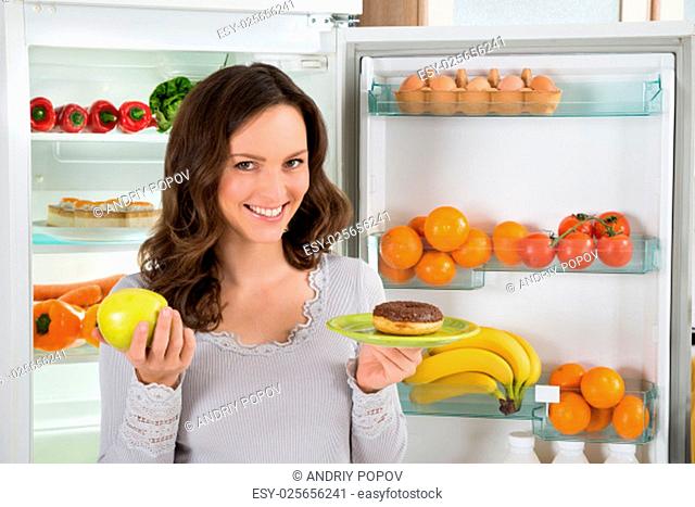 Young Happy Woman Holding Green Apple And Donut In Front Of Fridge