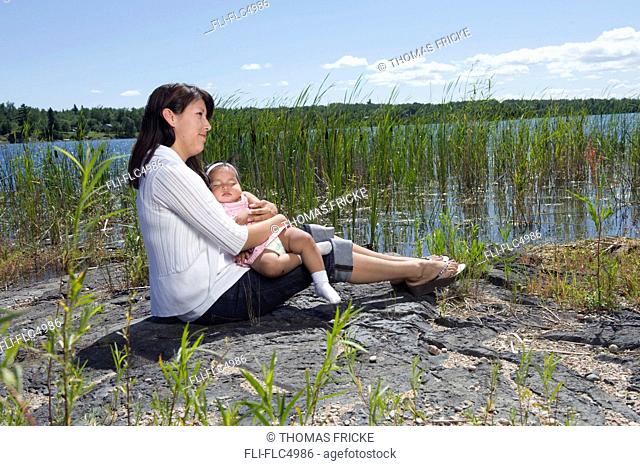Native aboriginal mother sitting with her one year old daughter on a rock beside a lake with reeds in Shoal Lake, Ontario, Canada