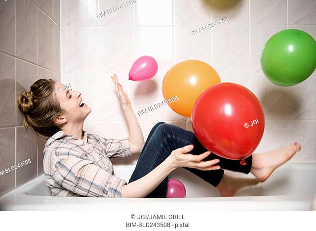 Laughing woman sitting in bathtub playing with multicolor balloons
