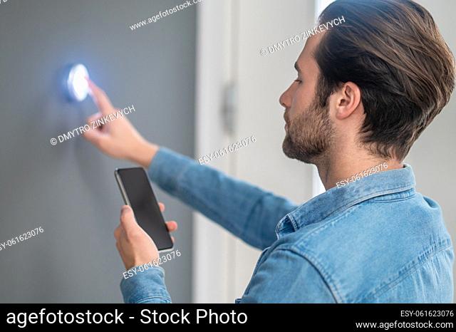 Home control panel. Focused young bearded man with smartphone touching with finger button on wall in new house