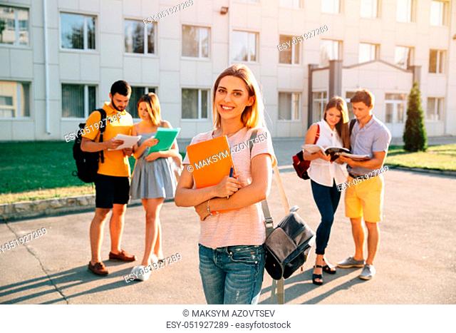 Student. Enjoying university life. Handsome young girl with grey velvet backpack holding books and smiling while standing against university with her friends in...
