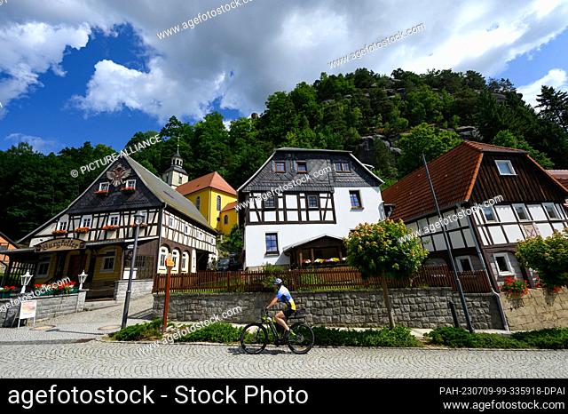 PRODUCTION - 06 July 2023, Saxony, Oybin: A cyclist rides past the Evangelical Lutheran Mountain Church below the fortress and the Oybin Monastery in the spa...