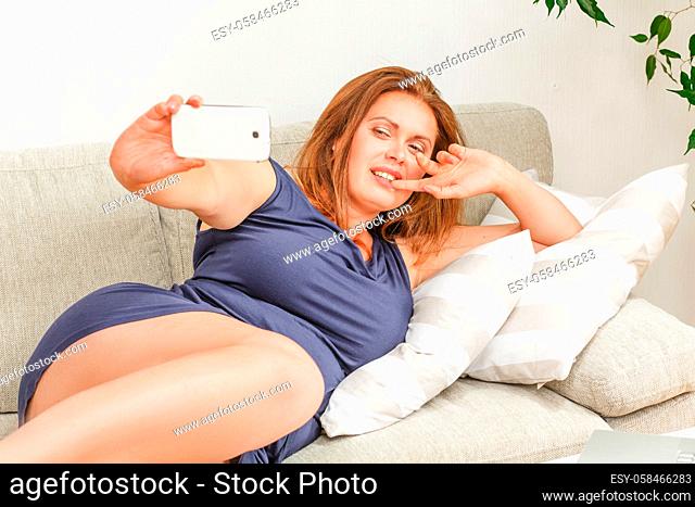 Closeup portrait of beautiful woman making selfies while lying on sofa or couch at home. Red haired lady showing yo sign to camera