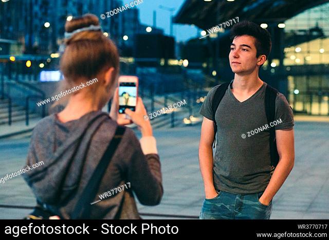 Young woman taking photos her friend, using a smartphone in the city at night