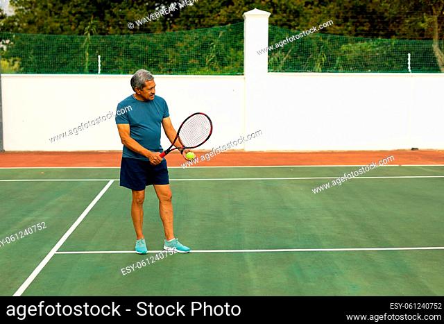 Full length of biracial senior man holding racket and ball playing tennis at tennis court